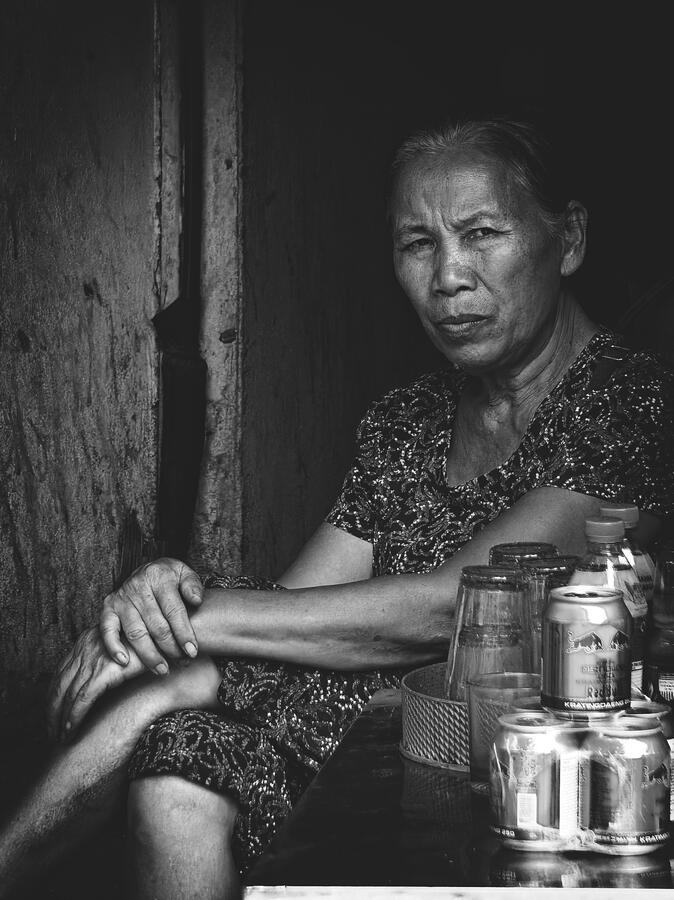 A woman sits quietly in a shaded doorway, her gaze meeting the camera. Captured in black and white, the portrait reveals the textures, expressions and quiet dignity of everyday life in Vietnam.