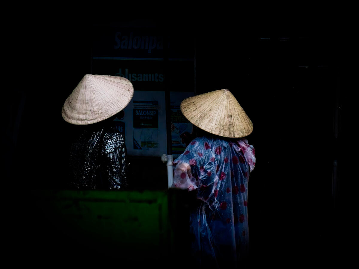 Two figures wearing traditional Vietnamese conical hats emerge from the darkness, their clothing and silhouettes illuminated by the available light. A quiet, unscripted moment of everyday life in Vietnam.