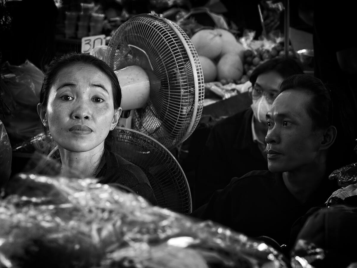 Black and white photograph of three market workers surrounded by produce and electric fans in Vientiane, Laos.