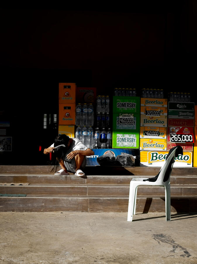 Woman crouching in strong sunlight outside a shop surrounded by Beerlao crates and an empty plastic chair in Vientiane, Laos.