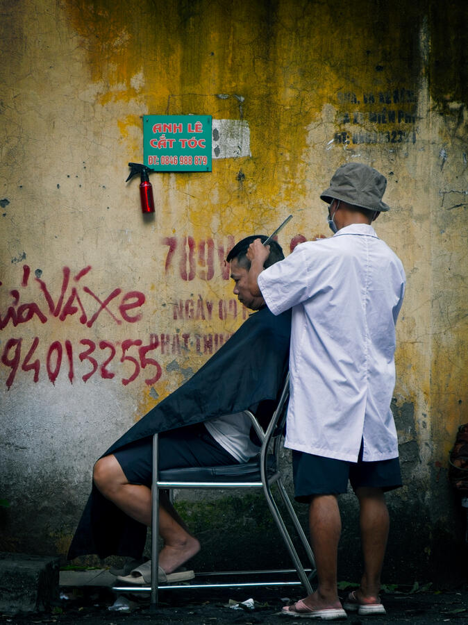 Street barber cutting a man's hair beside a weathered yellow wall in Hanoi, Vietnam.