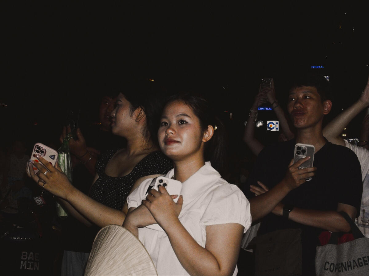 A crowd gathers beneath the night sky in Da Nang, Vietnam, their faces illuminated by the camera's flash. Surrounded by people capturing the occasion on their phones, a fleeting moment of anticipation becomes a portrait of modern street life.