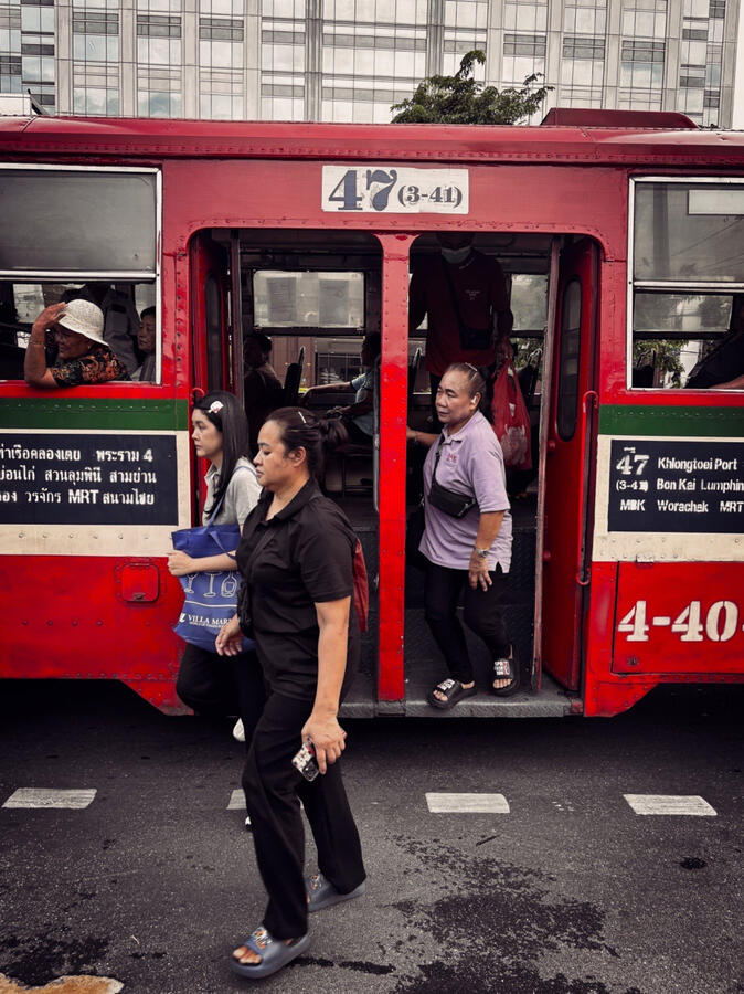 Passengers getting off a red city bus as pedestrians cross the street in Bangkok, Thailand.